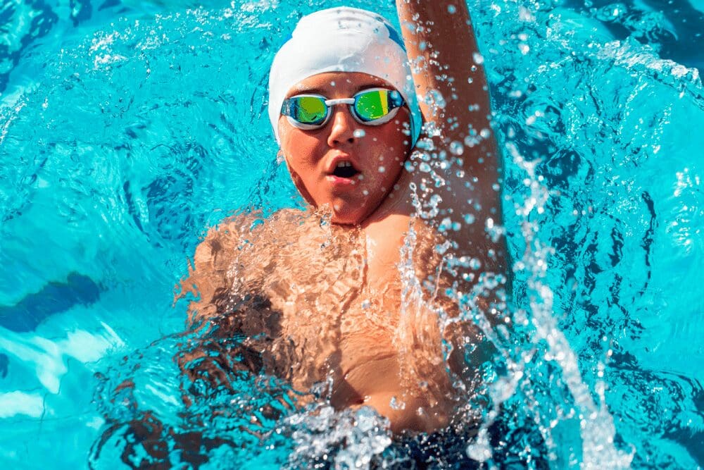 Person wearing a swim cap and goggles is swimming in a pool, splashing water as they move.