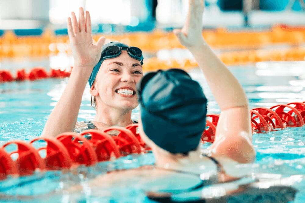 Two swimmers joyfully share a high-five near a lane divider in the pool, each wearing Quisque Rutrum swim caps and goggles, smiling broadly in the water.