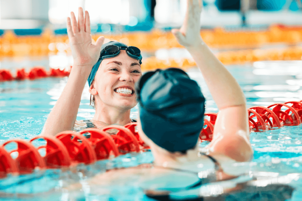 Two swimmers joyfully share a high-five near a lane divider in the pool, each wearing Quisque Rutrum swim caps and goggles, smiling broadly in the water.