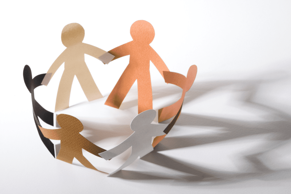 Paper cutouts of diverse human figures holding hands in a circle, casting shadows on a white surface.