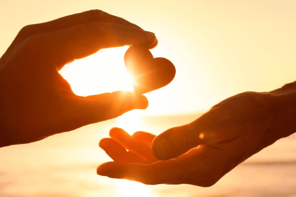 Two hands exchange a small heart-shaped object against a glowing sunset backdrop.