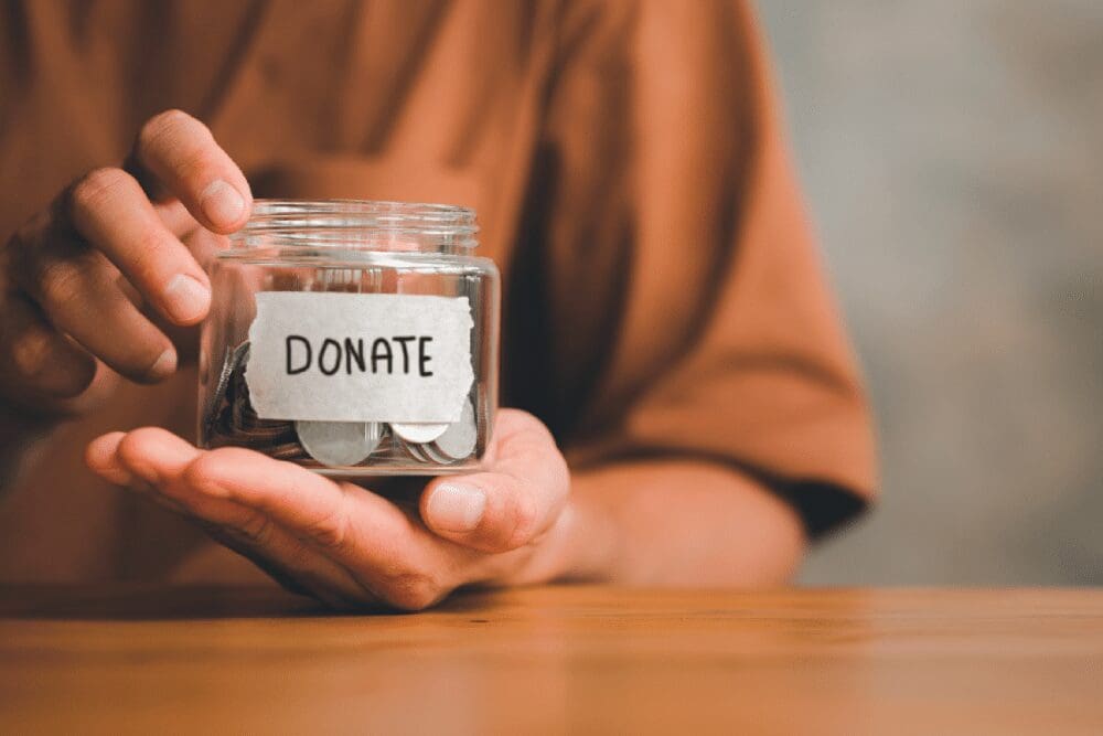 A person holds a glass jar labeled "Donate," containing coins, on a wooden surface.