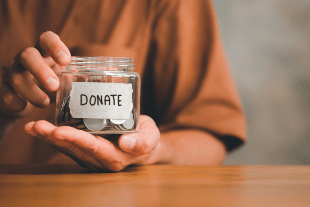 A person holds a glass jar labeled "Donate," containing coins, on a wooden surface.