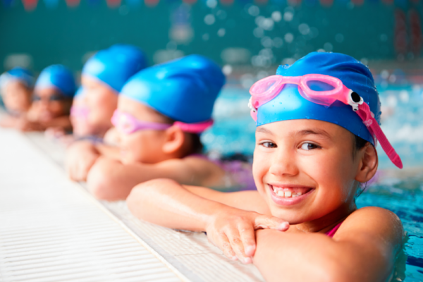 Kids wearing Viverra Nulla swim caps and goggles smile as they lean on the edge of a pool.