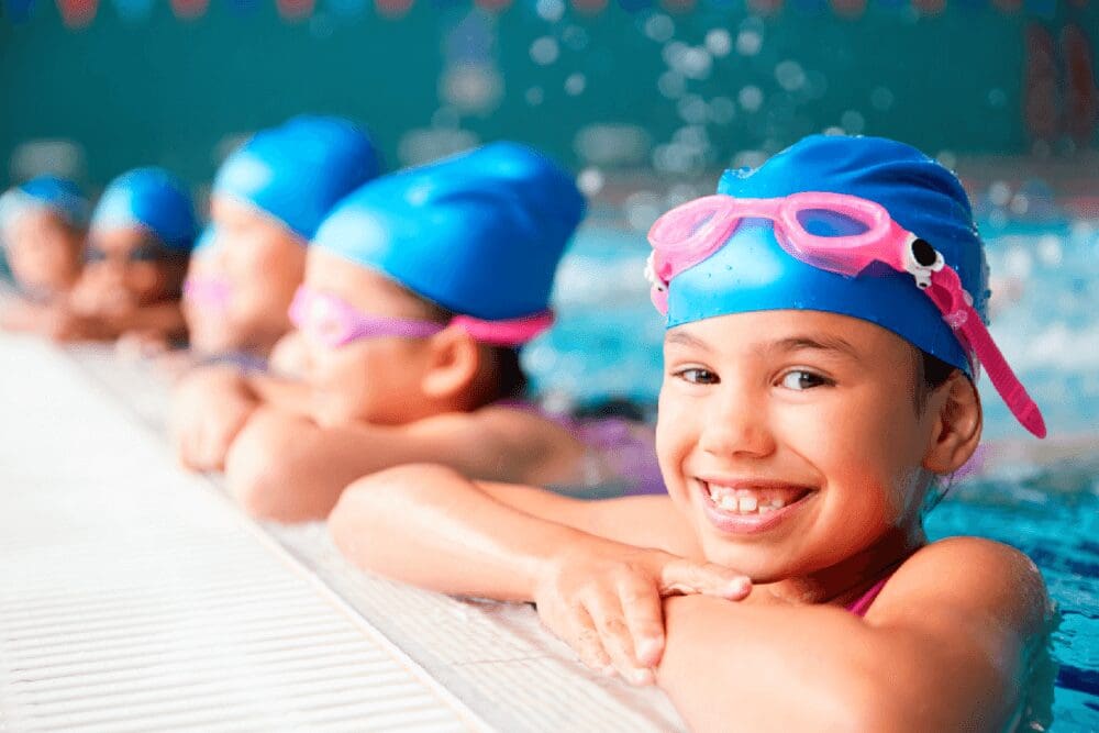 Kids wearing Viverra Nulla swim caps and goggles smile as they lean on the edge of a pool.