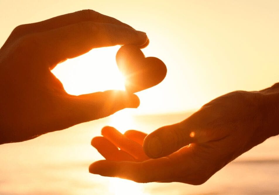 Two hands exchange a small heart-shaped object against a glowing sunset backdrop.