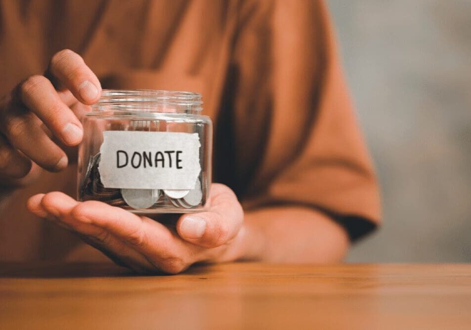 A person holds a glass jar labeled "Donate," containing coins, on a wooden surface.